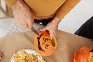 Close up of man hands carving Jack O Lattern from ripe orange pumpkin with knife on her wooden kitchen table. male preparing all hallows eve Halloween party decorations. Background, copy space. photo