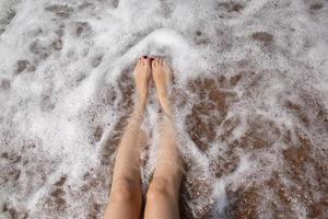 concepto de viaje - piernas de mujer en una hermosa playa tropical con arena de guijarros. pies sobre arena y olas en verano. chica descalza en el agua del océano en viajes de vacaciones. siéntete feliz y relájate. copie el espacio foto