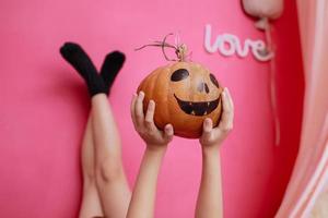 cropped photo of little girl legs and hands with Halloween pumpkin lying on bed in her pink room. Halloween party at home