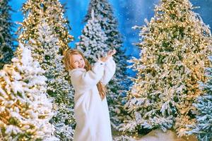 Linda chica rubia caucásica en abrigo de piel ecológica blanca caminando en el bosque de Navidad de invierno con luces. año nuevo, concepto de cuento de hadas foto