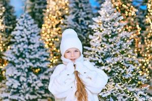 cute caucasian blonde girl in white eco fur coat, hat and gloves walking in the winter christmas forest. New year, fairy tale, fashion concept photo