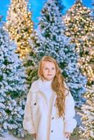 cute caucasian blonde girl in white eco fur coat walking in winter christmas forest with lights. New year, fairy tale concept photo