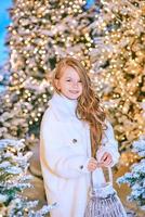 Linda chica rubia caucásica en abrigo de piel ecológica blanca caminando en el bosque de Navidad de invierno con luces. año nuevo, concepto de cuento de hadas foto