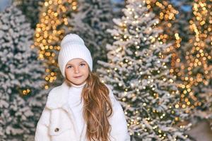 cute caucasian blonde girl in white eco fur coat, hat and gloves walking in the winter christmas forest. New year, fairy tale, fashion concept photo