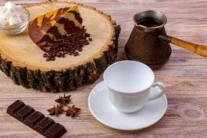 A coffee cup with a piece of cake on wooden stump, a chocolate bar, anise, coffee beans and bowl with sugar cubes on a wooden table photo