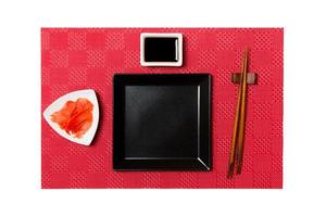 Empty black square plate with chopsticks for sushi and soy sauce, ginger on red mat sushi background. Top view with copy space for you design photo