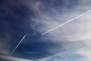 Aircraft condensation contrails in the blue sky inbetween some clouds photo