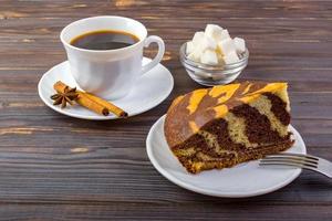 A tasty cake and a cup of coffee. A bowel with sugar cubes and cinnamon sticks on a small plate near the coffee and a fork on dark wooden background photo