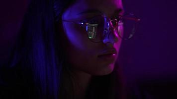 Young woman in a dark purple room with laptop screen reflection on eyeglasses video