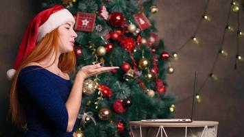 een mooi roodharig vrouw in een de kerstman hoed feliciteert familieleden Aan Kerstmis door videoconferentie Aan een laptop. afgelegen communicatie in de nieuw jaar ten gevolge naar coronavirus. modern technisch concept. video