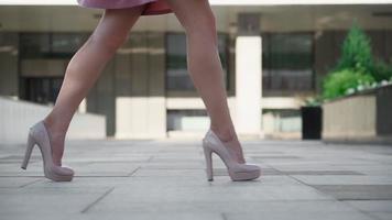 sexy riuscito attività commerciale donna nel un' rosa vestito a piedi nel il città parco. bellissimo femmina gambe nel alto tacchi siamo a piedi lungo un' urbano strada contro il fondale di tramonto luce. lento movimento. video