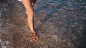 female legs are walking along the beach along the sea. The woman walks barefoot splashing in the sea water. Summer vacation and relaxation. Slow motion.