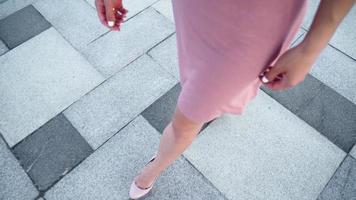 sexy erfolgreiche Geschäftsfrau in einem rosafarbenen Kleid, das im Stadtpark spazieren geht. schöne weibliche beine in high heels gehen vor dem hintergrund des sonnenuntergangslichts eine städtische straße entlang. Zeitlupe. video