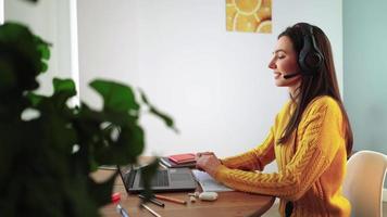una mujer feliz con auriculares participa en el aprendizaje electrónico por chat de cámara web en casa. una joven estudiante con suéter amarillo en una laptop se comunica en línea por videollamada. educación a distancia y concepto de tecnología moderna.