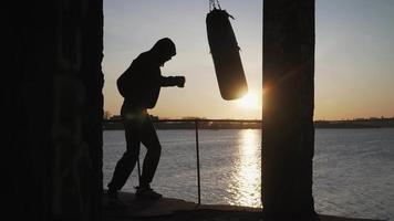 el boxeador recortado contra el telón de fondo de una hermosa puesta de sol golpea un saco de boxeo rápidamente hasta el fracaso. fatiga, ya no puede hacer ejercicio. motivación. deportes y artes marciales. video