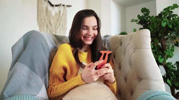 Relaxed Woman in Yellow Sweater Smiling Hold Smartphone Watching Social Media Stories Video Sit on Sofa Home. Happy Girl Look Cellphone Laughing Enjoying Using Mobile App Having Fun Play and Chatting