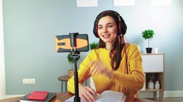 glückliche junge lehrerin in kopfhörern kommuniziert per videoanruf mit handy. Frau im gelben Pullover lächelt und spricht online, während sie zu Hause am Tisch sitzt. Fernunterricht und E-Learning-Konzept video