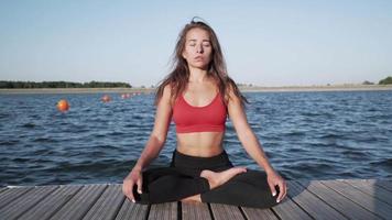 Young athletic Caucasian girl in a red tank top practices yoga on a lake background. Sits in a lotus position on a pantone. video