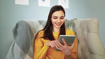 Young happy woman in yellow sweater sitting on cozy sofa at home smiles and studies app on digital computer tablet. Beautiful girl in good mood uses portable device to communicate on social networks. video
