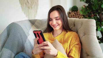 entspannte frau im gelben pullover, die lächelnd das smartphone hält, das social-media-geschichtenvideo anschaut, sitzt auf dem sofa zu hause. glückliches Mädchen sieht Handy lachen und genießt die Verwendung der mobilen App, die Spaß beim Spielen und Chatten hat