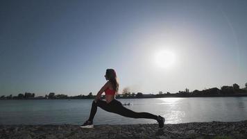 A young athletic Caucasian girl in a red tank top and leggings is doing stretching on the background of the lake. Flexibility. Learning to do split. video