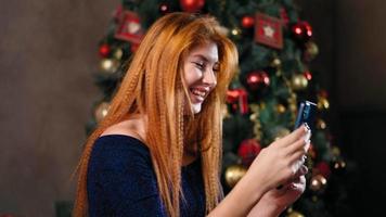 A beautiful red-haired woman sits next to a Christmas tree decorated with flickering lights and uses her smartphone to communicate remotely. Listens to New Year's greetings, laughs and is happy. video