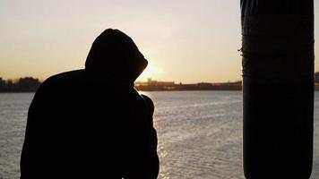 silhouet van een bokser tegen de backdrop van een mooi zonsondergang en een rivier- in een verlaten gebouw, treinen slagen Aan een ponsen tas. episch video. motivatie. sport- en krijgshaftig kunsten. langzaam beweging. video