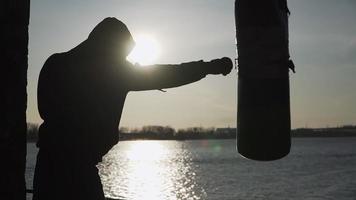 Silhouette of a boxer against the backdrop of a beautiful sunset and a river in an abandoned building, trains blows on a punching bag. Epic video. Motivation. Sports and martial arts. Slow motion. video