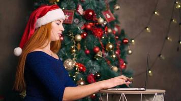 una hermosa mujer pelirroja con un sombrero de santa trabaja en una laptop y escribe en un teclado contra el fondo de un árbol de navidad. material de archivo 4k de alta calidad video