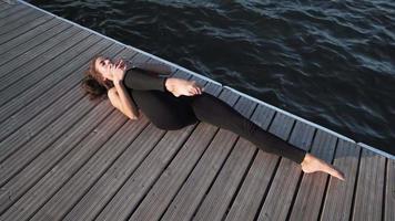 jeune fille caucasienne athlétique dans un débardeur rouge et des leggings fait des étirements et du yoga sur le fond du lac. souplesse. video