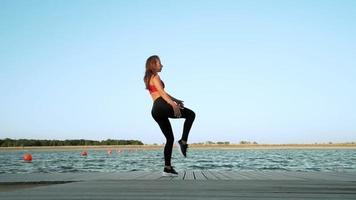 ein junges athletisches kaukasisches mädchen in einem roten tanktop und leggings zeigt ihre dehnbarkeit und flexibilität vor dem hintergrund eines sees. schöner Tanz. dehnen, spalten. video
