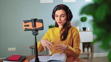 Happy young teacher in headphones communicates by video call using mobile phone. Woman in yellow sweater smiles and talks online while sitting table at home. Distance education and e learning concept