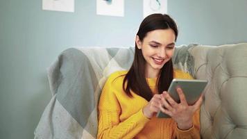 Young happy woman in yellow sweater sitting on cozy sofa at home smiles and studies app on digital computer tablet. Beautiful girl in good mood uses portable device to communicate on social networks. video