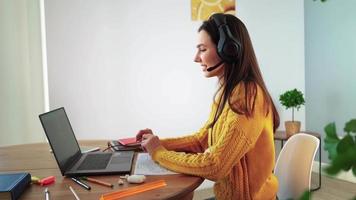 mulher feliz em fones de ouvido está engajada e aprendendo pelo bate-papo da webcam em casa. jovem estudante de suéter amarelo no laptop se comunica on-line por videochamada. educação a distância e conceito de tecnologia moderna. video