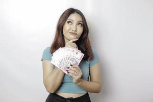 una joven pensativa lleva una camiseta azul y tiene dinero en efectivo en rupias indonesias aislada de fondo blanco foto