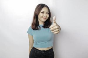 Excited Asian woman wearing blue shirt gives thumbs up hand gesture of approval, isolated by white background photo