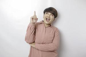 Excited Asian woman wearing pink shirt pointing at the copy space upside her, isolated by white background photo