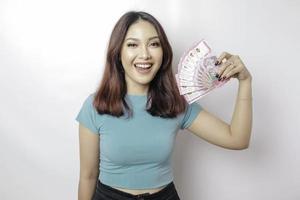 una joven feliz lleva una camiseta azul y tiene dinero en efectivo en rupias indonesias aislada de fondo blanco foto