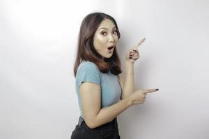 Shocked Asian woman wearing blue t-shirt pointing at the copy space beside her, isolated by white background photo