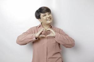 An attractive young short-haired Asian woman wearing a pink shirt feels happy and a romantic shapes heart gesture expresses tender feelings photo