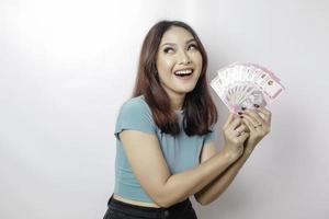 una joven feliz lleva una camiseta azul y tiene dinero en efectivo en rupias indonesias aislada de fondo blanco foto