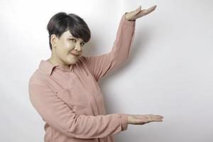 Excited short-haired Asian woman wearing a pink shirt pointing at the copy space beside her, isolated by a white background photo