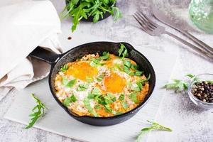 Shakshuka with poached eggs in spicy tomato pepper sauce and arugula in a frying pan on the table