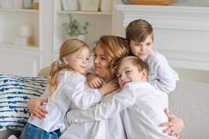 te amamos. mujer joven sonriente, los niños abrazan a mamá mientras se sientan en el sofá en la vida foto