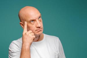 portrait of a bald man in a white t-shirt in the background, pensive expression, holding a finger near his temple photo