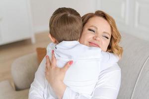 Happy cute caucasian little son hugs mom while sitting on sofa in living room photo