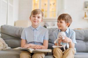 Two boys sitting on couch are reading book. Older brother reads interesting photo