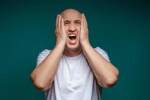 Bald man holds his hands up to his face in horror against a blue background photo