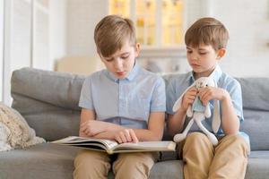 Two boys sitting on couch are reading book. Older brother reads interesting photo