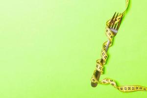 Top view composition of fork wrapped with measuring tape on green background with empty space for your ideas. Overweight and overeating concept photo